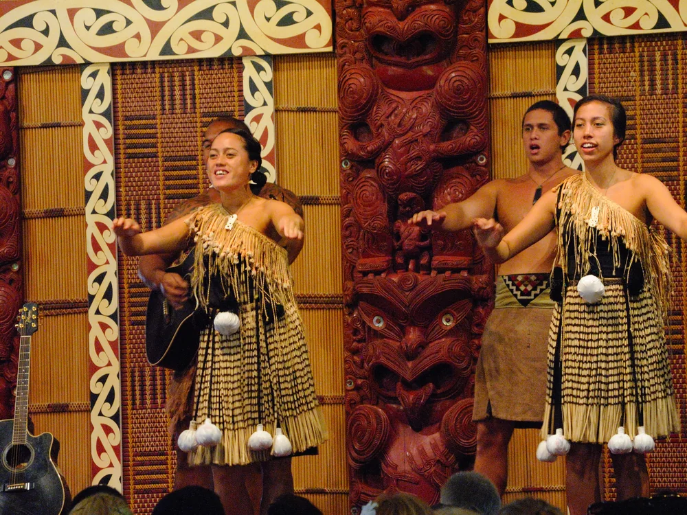 Māori performers in traditional dress dancing in front of a carved meeting-house façade at the Polynesian Cultural Center's Aotearoa village